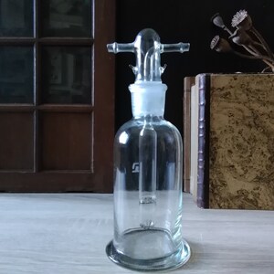 May include: A clear glass bottle with a glass stopper and a glass tube extending from the stopper to the bottom of the bottle. The bottle has a round base and a cylindrical body. The bottle is sitting on a wooden surface.