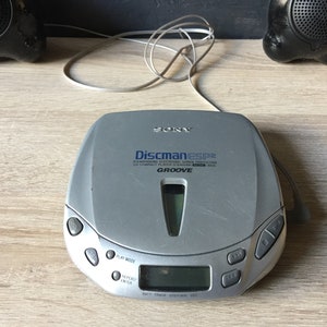 Può includere: Un lettore CD portatile Sony Discman ESP2R argentato con un display nero e blu e pulsanti. Il lettore ha il testo "Discman ESP2R" e "GROOVE" stampato su di esso.