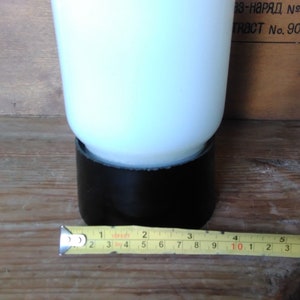 May include: A white opaque glass object with a black base, possibly a vase or lamp. A yellow measuring tape shows the object's base is approximately 2 inches wide. The background is a wooden surface.