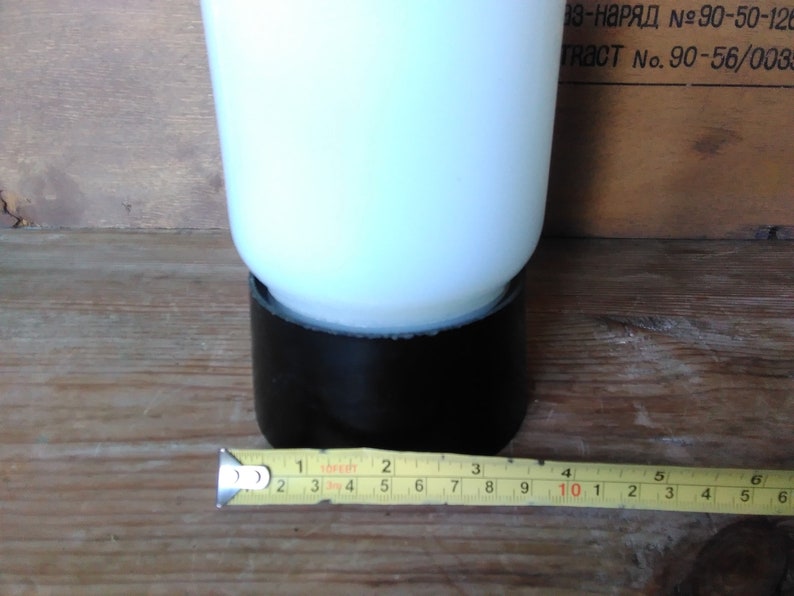 May include: A white opaque glass object with a black base, possibly a vase or lamp. A yellow measuring tape shows the object's base is approximately 2 inches wide. The background is a wooden surface.