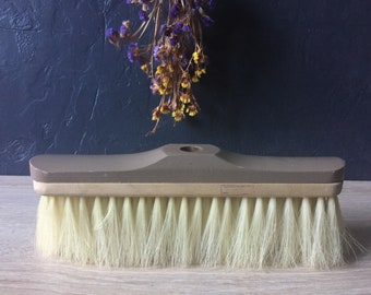 May include: A brown wooden-handled broom with a long, white bristle brush. The broom is on a light wood surface.
