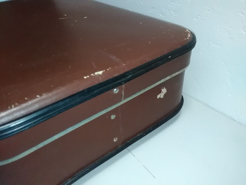 Może przedstawiać: Brązowa vintage'owa walizka z czarną obw&oacute;dką i srebrnym akcentem. Walizka nosi ślady zużycia, co nadaje jej vintage'owy wygląd.