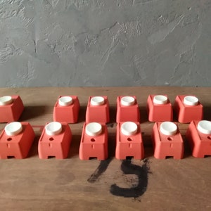 May include: A collection of 13 red plastic push buttons with white centers. The buttons are arranged in two rows on a wooden surface.