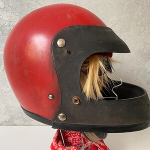 May include: A vintage red motorcycle helmet featuring a black visor and a black chin guard. The helmet has a smooth, rounded shell with visible surface wear and is mounted on a stand with a red patterned fabric visible at the base.