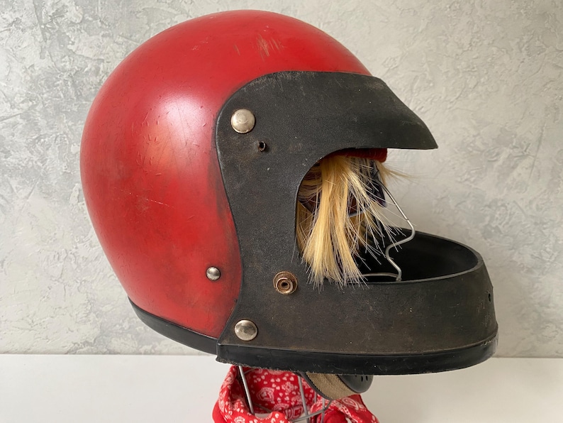 May include: A vintage red motorcycle helmet featuring a black visor and a black chin guard. The helmet has a smooth, rounded shell with visible surface wear and is mounted on a stand with a red patterned fabric visible at the base.