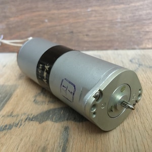 May include: A silver and black electric motor with a black label that reads "1-6M". The motor has a shaft with a small gear on the end.