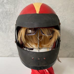 May include: A red and black full-face helmet featuring a yellow stripe on the top. Inside the helmet, a blonde wig is displayed on a metal stand, with a pair of dark-tinted sunglasses positioned over the hair, visible through the open visor.
