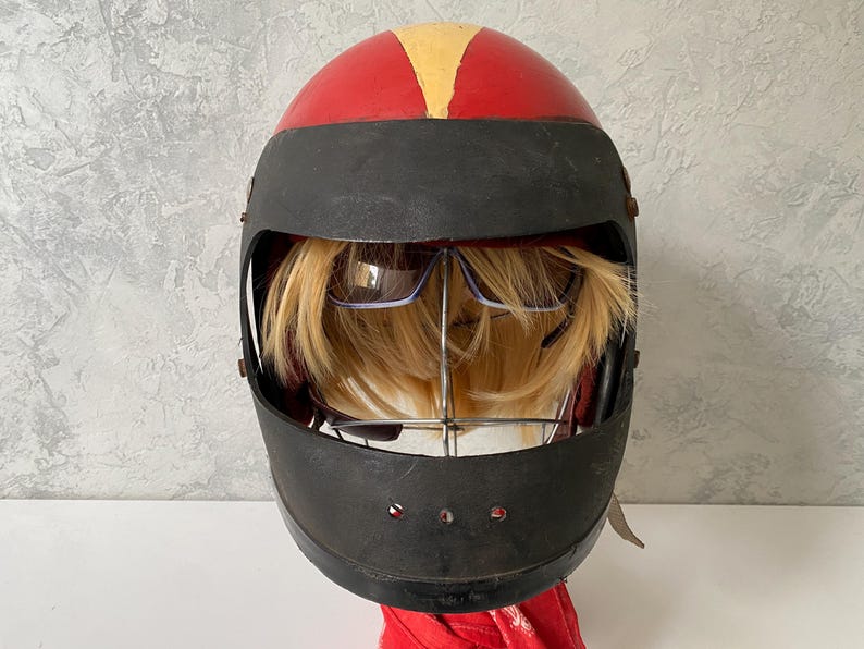 May include: A red and black full-face helmet featuring a yellow stripe on the top. Inside the helmet, a blonde wig is displayed on a metal stand, with a pair of dark-tinted sunglasses positioned over the hair, visible through the open visor.