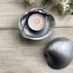 May include: A silver-painted seashell candle holder with a lit tea light candle inside. Another silver seashell is next to the candle holder. The items are on a light grey wooden surface. Decorative home decor.