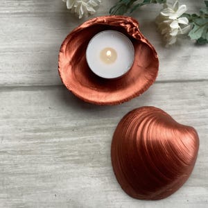 May include: A copper-coloured seashell candle holder with a lit white tea light candle. The holder is made from a large seashell, with a matching shell next to it. Decorative flowers are in the background.