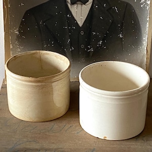 Könnte beinhalten: Zwei antike Keramikgefäße mit cremefarbener Glasur stehen auf einer Holzoberfläche. Die Gefäße sind zylindrisch mit leicht ausgestelltem Rand. Dahinter befindet sich ein Vintage-Schwarz-Weiß-Porträt eines Mannes im Anzug.