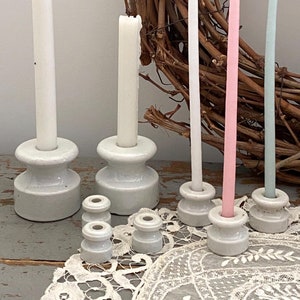 Op de afbeelding: Vijf witte keramische kaarsenstandaards met vier witte kaarsen en één roze en één blauwe kaars. De kaarsen zijn in verschillende maten en zijn gerangschikt op een wit kantkleed.