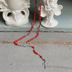 Può includere: Un rosario rosso e dorato con una croce d'argento. Il rosario è appoggiato su una superficie di legno con due vasi di porcellana bianca sullo sfondo.