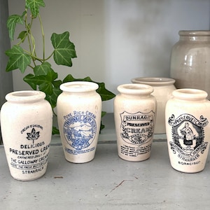 Pode incluir: Quatro potes de creme de cerâmica em tom esbranquiçado com corpos cilíndricos e gargalos estreitos. Cada pote apresenta textos e ilustrações impressos em preto ou azul, incluindo rótulos como Strathbogie Dairy, Dunragit e Wigtownshire Creamery.