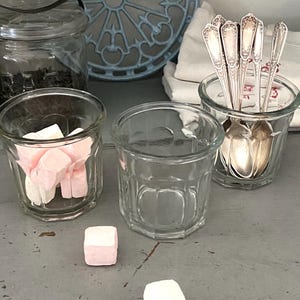 以下が含まれることがあります： 透明なガラス瓶とカップのコレクション。マシュマロと銀食器が入っているものもあります。背景には青い装飾的な金属片があります。瓶は灰色の表面に配置されており、ヴィンテージな美しさを生み出しています。