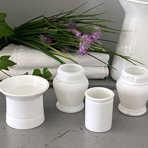 Pode incluir: Uma coleção de recipientes de cerâmica branca de várias formas, incluindo um pequeno jarro, dispostos sobre uma superfície cinza. Um maço de cebolinho com flores roxas e toalhas brancas ao fundo.