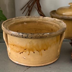 May include: A round, low-profile ceramic bowl with a mustard-yellow glaze and a darker, textured rim. The bowl features two small, integrated side handles and a visible crackled finish across the surface, with darker brown drip-like markings.