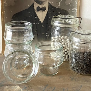 Peut inclure: Plusieurs bocaux en verre transparent de différentes tailles, certains avec des couvercles et des fermetures métalliques, sont disposés sur une surface en bois. Un bocal contient de petites sphères blanches, un autre des feuilles séchées. Un portrait vintage en noir et blanc est en arrière-plan.