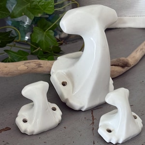 Op de afbeelding: Drie witte keramische wandhaken met een gladde, glanzende afwerking. De haken hebben een gebogen, T-vormig ontwerp met een brede basis en twee voorgeboorde schroefgaten aan elke kant voor montage. De set bevat één grote haak en twee kleinere haken.