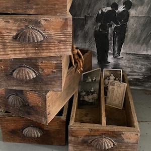 Può includere: Una pila di scatole di legno consumate con maniglie a forma di conchiglia. Le scatole sono di varie dimensioni e mostrano segni di età. Alcune scatole contengono vecchie fotografie e una figura di artista in legno. Un disegno a carboncino è sullo sfondo.