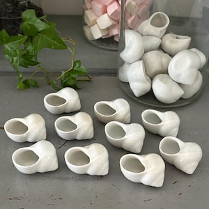 Peut inclure: Une collection de petits objets en céramique blanche brillante en forme de coquillage disposés sur une surface grise. Plusieurs de ces répliques de coquillages creux à motifs en spirale sont éparpillées au premier plan, d'autres dans un bocal.
