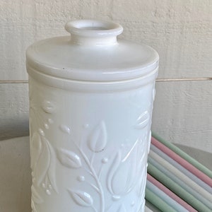 Könnte beinhalten: Ein weißes, zylindrisches Glas mit Deckel und floralem Reliefdesign. Das Glas besteht aus undurchsichtigem Glas und hat eine glatte, glänzende Oberfläche. Eine Sammlung pastellfarbener Kerzen befindet sich neben dem Glas.