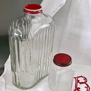 Peut inclure: Deux bocaux en verre transparent avec des couvercles rouges. Le plus grand bocal a des nervures verticales et le plus petit bocal a une surface texturée. Les deux bocaux sont posés sur un tissu blanc.