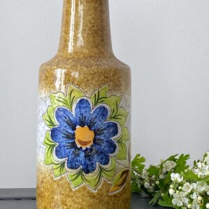Peut inclure: Un vase en céramique avec une finition marron tachetée et texturée. Une grande fleur bleue avec un centre jaune et des feuilles vertes est peinte sur le devant. Le vase a un col long et fin et un corps arrondi.