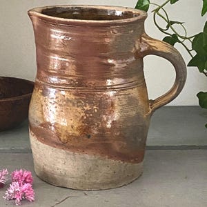 Peut inclure: Une cruche en céramique rustique et faite à la main avec une anse incurvée. La cruche présente un design bicolore avec une base beige clair et une section supérieure marron. La surface a une finition texturée et vernissée, et la cruche mesure environ 20 cm de haut.