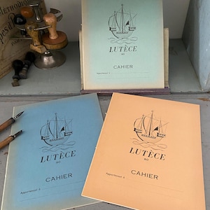 Può includere: Quaderni in azzurro, pesca e verde menta con un'illustrazione di una nave e la parola "Lutèce" stampata sulla copertina. La parola "Cahier" è stampata anche sulla copertina. Altri articoli di cancelleria sullo sfondo.