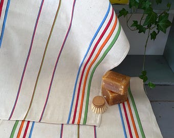 Puede incluir: Dos toallas de cocina blancas con rayas de colores en rojo, naranja, verde y azul. Las toallas están dobladas y colocadas sobre una superficie de madera. Hay dos barras de jabón y un cepillo de madera encima de las toallas.