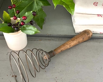 Può includere: Una frusta a filo d'epoca con manico in legno è appoggiata su una superficie grigia usurata. Le spirali metalliche della frusta sono avvolte strettamente. Un piccolo vaso di ceramica bianca con bacche e foglie verdi è sullo sfondo.