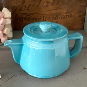 May include: A small, turquoise ceramic teapot with a lid and handle. The teapot has a spout for pouring and is set against a blurred background. The teapot is a solid color and appears to be vintage.