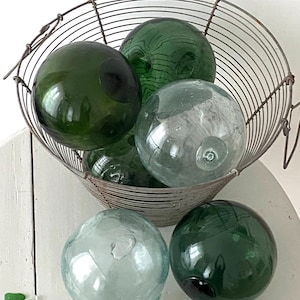 Può includere: Una collezione di galleggianti da pesca in vetro verde e trasparente in un cesto di filo metallico. I galleggianti sono rotondi e hanno una superficie liscia e lucida.