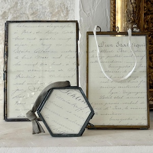 Könnte beinhalten: Zwei rechteckige Glasrahmen mit dunklen Metallkanten und ein sechseckiger Glasrahmen mit dunklem Metallrand. Jeder Rahmen enthält cremefarbenes Papier mit handschriftlicher französischer Schreibschrift. Der sechseckige Rahmen hat eine graue Stoffschlaufe.