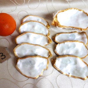 Pode incluir: Um conjunto de pratos em forma de concha de ostra branca com bordas douradas, dispostos sobre uma superfície estampada. Uma fruta laranja e uma pequena etiqueta com o número "3" também estão presentes. Os pratos têm um brilho perolado.