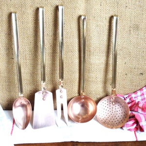 Op de afbeelding: Een set van vijf koperen keukengerei met lange metalen handvatten, waaronder een massieve lepel, een platte spatel, een vork met twee tanden, een ronde soeplepel en een schuimspaan, gerangschikt op een witte doek met een rood geruite servet.
