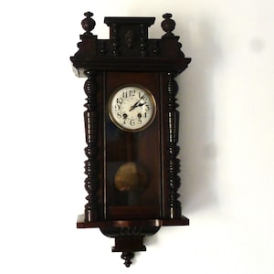 Può includere: Un orologio da parete antico in legno scuro con un quadrante bianco e indici orari romani neri. L'orologio ha un pendolo e una parte superiore decorativa.