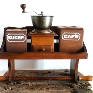 Puede incluir: Un soporte de pared de madera para café con un molinillo de café de manivela, un tarro de azúcar etiquetado como "SUCRE" y un tarro de café etiquetado como "CAFE".