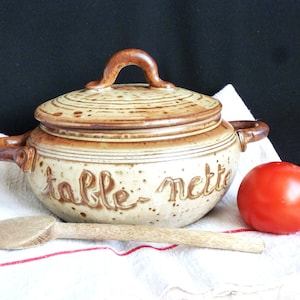 Peut inclure: Un plat de service en céramique brun avec un couvercle et deux poignées. Le plat porte l'inscription "table nette" sur le côté. Une cuillère en bois et une tomate rouge sont également présents dans l'image.