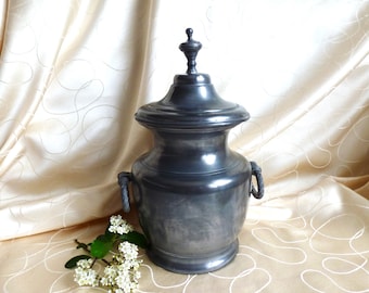 May include: A pewter urn with a lid and two handles. The urn is a dark gray color and has a smooth, polished finish. The lid has a finial on top. The urn is sitting on a cream-colored fabric background.