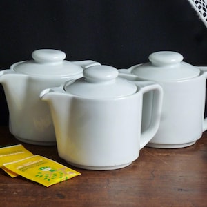 May include: Three white ceramic teapots with lids. The teapots are arranged in a row on a wooden surface.