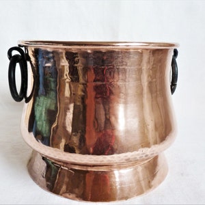 Könnte beinhalten: Ein Kupferkessel mit gehämmertem Finish. Der Topf hat einen abgerundeten Körper, einen ausgestellten Boden und zwei schwarze Metallgriffe. Das Kupfer hat einen warmen, reflektierenden Glanz. Dieses dekorative Stück eignet sich für die Inneneinrichtung.