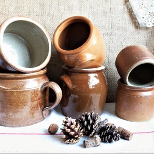 Puede incluir: Cinco ollas de cerámica marrón con asas, algunas con tapas, dispuestas sobre una superficie de lino blanco con piñas y bellotas.