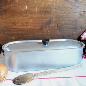 Op de afbeelding: Een zilveren ovale aluminium stoompan met een zwarte handgreep en een zwarte knop op de deksel. De stoompan staat op een wit tafelkleed met een houten lepel en een bruine ui.