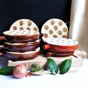 Puede incluir: Un conjunto de cazuelas de cerámica marrón para caracoles con asas, junto con dos platos de servicio a juego. Las cazuelas están sobre una tabla de madera, con hierbas frescas y una cebolla en primer plano. El fondo es oscuro.