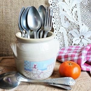 Puede incluir: Un tarro de cerámica beige lleno de cucharas y un tenedor de acero inoxidable, con una calcomanía de una persona sentada en una silla junto al mar. Una naranja fresca y dos cucharas plateadas con mangos florales descansan cerca.