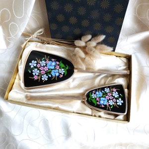 May include: A vintage hand mirror set with two gold-trimmed mirrors featuring a floral design with blue and pink flowers on a black background. The mirrors are in a white satin-lined box.