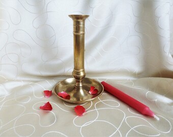May include: A brass candlestick with a round base and a tall, cylindrical stem. The candlestick is on a white surface with red rose petals scattered around it. A red candle is lying next to the candlestick.