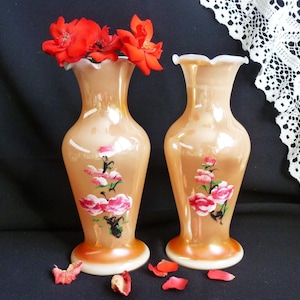 Może przedstawiać: Dwa brzoskwiniowe wazony z falbankami, każdy ozdobiony kwiatowym wzorem z różowych róż i zielonych liści. Wazony zawierają żywe czerwone róże. Wazony stoją na ciemnej powierzchni.